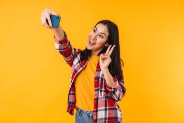 Sarı arka planda izole edilmiş cep telefonuyla selfie çeken hoş bir kadının fotoğrafı.