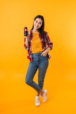 Sarı arka planda izole edilmiş gülümseyen ve soda içen neşeli bir kadının fotoğrafı.