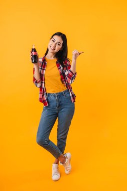 Neşeli, hoş bir kadının işaret ettiği ve sarı arka planda izole edilmiş gazoz içtiği fotoğraf.