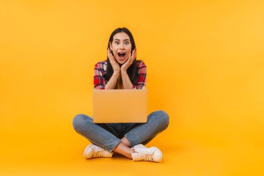 Sarı arka planda dizüstü bilgisayarla yerde otururken şaşkınlığını ifade eden güzel bir kadının fotoğrafı.