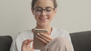 Gözlüklü neşeli esmer kadın evde kanepede otururken akıllı telefon kullanıyor.
