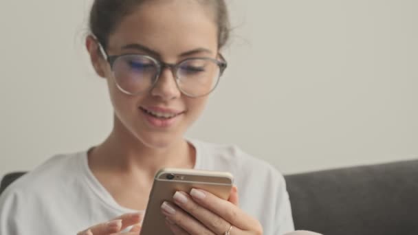 Sorrindo Mulher Morena Bonita Óculos Usando Smartphone Enquanto Sentado Sofá — Vídeo de Stock