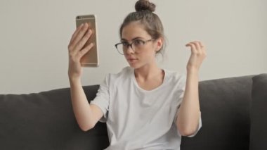 Gözlüklü güzel esmer kadın ayna gibi akıllı telefon kullanıyor ve evdeki kanepede otururken saçını düzeltiyor.