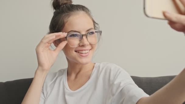 幸せなかわいいですブルネット女性で眼鏡作り自画撮りスマートフォン上の座っている間にソファに家 — ストック動画