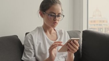 Gözlüklü esmer kadın akıllı telefon kullanıyor ve evde koltukta otururken mutlu oluyor.