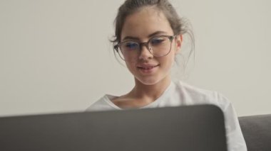 Gözlüklü mutlu esmer kadın evde kanepede otururken dizüstü bilgisayar kullanıyor.