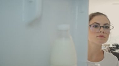 Gözlüklü, konsantre olmuş esmer kadın evdeki mutfaktaki buzdolabında bir şey arıyor.