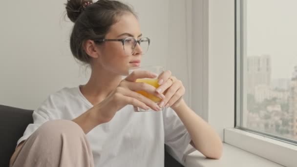 幸せなかわいいですブルネット女性で眼鏡飲みジュースと見て離れて座っています上のソファに家 — ストック動画