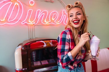 Eski Amerikan kafesinde müzik kutusuna yaslanırken güzel saç stiliyle milkshake içen neşeli genç bir kadının fotoğrafı.