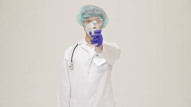 Genç Asyalı doktor, beyaz duvar arkasında steril üniformayla izole edilmiş. Vücut sıcaklığını temassız termometreyle ölçüyor.