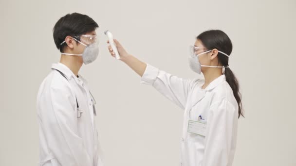 身穿无菌制服 手持无接触式温度计 头戴口罩 被隔离在白底之上的年轻亚洲医生测量体温 — 图库视频影像