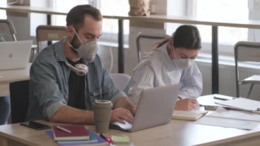 Genç konsantre iş adamları ofiste Coronavirus yüzünden maske takarak dizüstü bilgisayar kullanarak not tutuyorlar.