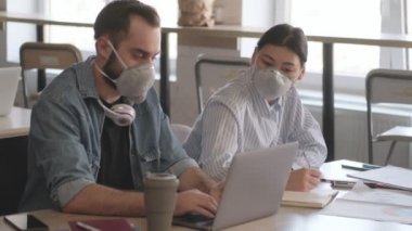 Genç, mutlu iş arkadaşları ofiste dizüstü bilgisayar kullanarak Coronavirus yüzünden maske takarak notlar yazıyorlar.