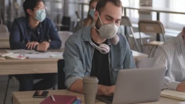 Genç memnun iş arkadaşları ofiste diz üstü bilgisayar kullanarak Coronavirus yüzünden maske takarak notlar yazıyorlar.