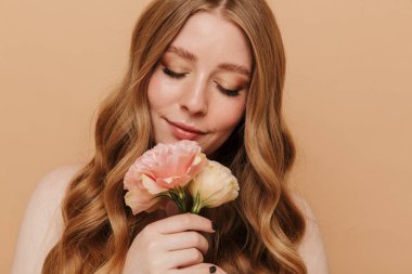 Bej arka planda saklanmış çiçekleri tutan, uzun kumral saçlı, muhteşem, yarı çıplak bir kadının portresi.