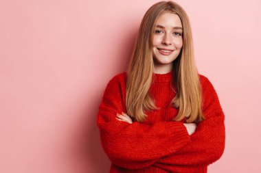 Kırmızı kazaklı mutlu bir kadının fotoğrafı. Elleri çaprazlanmış pembe duvarda poz verirken gülümsüyor.