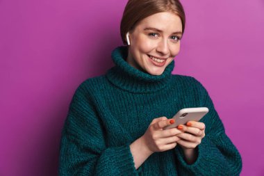 Yeşil kazaklı, gülümseyen beyaz bir kadının cep telefonu ve menekşe duvarda izole edilmiş kablosuz kulaklık kullandığını gösteren bir fotoğraf.