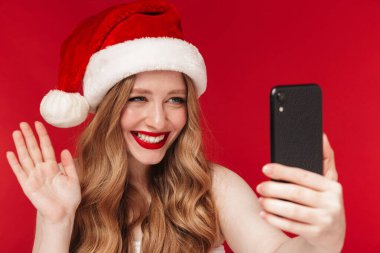 Kırmızı duvar arkasında izole edilmiş, parlak kırmızı ruj süren, Noel şapkası takan ve cep telefonuyla konuşan güzel mutlu bir kadının görüntüsü..