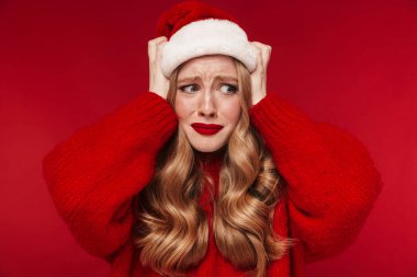 Kafası karışmış, kırmızı duvar arkasına izole edilmiş kırmızı rujlu, sıcak kırmızı kazak giyen ve Noel şapkası takan üzgün, beyaz bir kadın resmi..