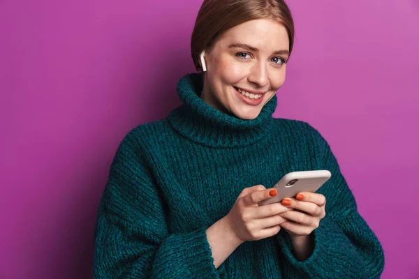 携帯電話と無線イヤフォンを使用して緑色のセーターで笑顔の白人女性の写真は 紫外線壁に隔離された — ストック写真