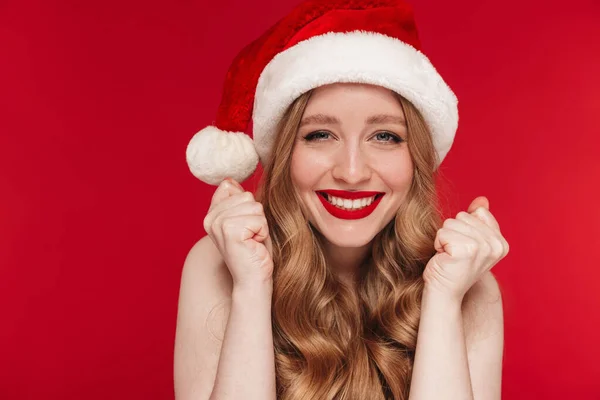 Image of a beautiful positive caucasian woman posing isolated over red wall background with bright red lipstick wearing christmas hat make winner gesture.