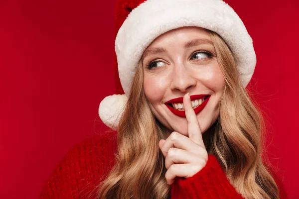 Image of a positive caucasian woman posing isolated over red wall background with bright red lipstick dressed in warm red sweater wearing christmas hat showing silence gesture.