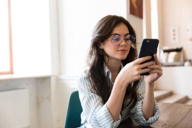 Kafe masasında oturan güzel, genç esmer kadın, cep telefonu kullanıyor.
