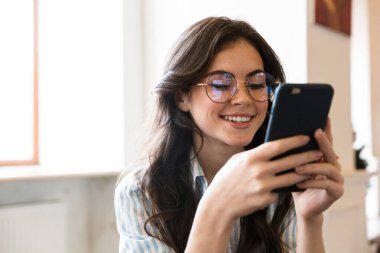 Güzel gülümseyen esmer kadın, kafeteryada oturuyor, cep telefonu kullanıyor.