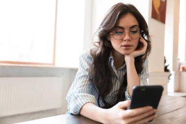 Kafe masasında oturan güzel, genç esmer kadın, cep telefonu kullanıyor.