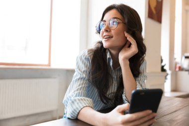 Güzel gülümseyen esmer kadın, kafeteryada oturuyor, cep telefonu kullanıyor.