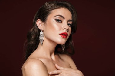 Küpe takan çekici, üstsüz bir kadın resmi. Burgonya arka planında izole edilmiş kameraya bakıyor.