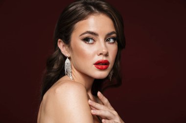 Küpe takan, burgonya rengi arka planda izole edilmiş, poz veren ve bir kenara bakan şık gömlekli bir kadın resmi.