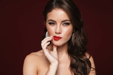 Kafası karışmış, küpeli, poz veren ve şarap rengi arka planda izole edilmiş kameraya bakan bir kadın resmi.