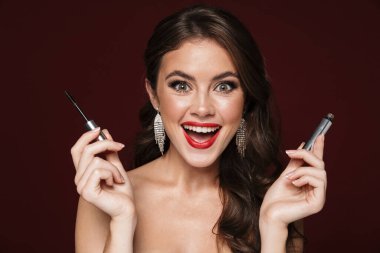 Heyecanlı, üstsüz, küpe takan, gülümseyen ve maskara tutan bir kadının resmi.