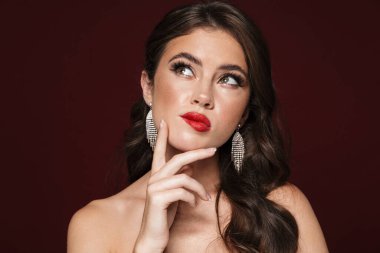 Küpe takan, poz veren ve yukarı bakan, burgonya arka planda soyutlanmış, üstsüz bir kadın görüntüsü.