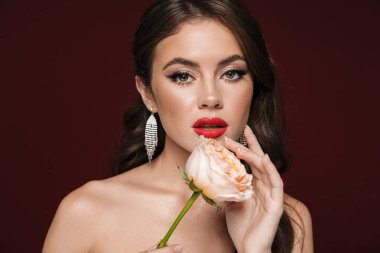 Küpe takan, Rose 'u tutan ve Burgonya arka planında izole edilmiş kameraya bakan romantik üstsüz bir kadın resmi.