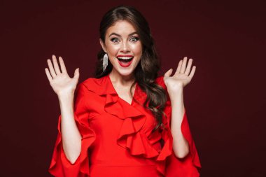 Kırmızı elbise giyen, gülümseyen ve şarap rengi arka planda izole edilmiş kameraya bakan şaşırmış bir kadın resmi.
