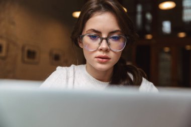 Kafede gümüş dizüstü bilgisayar kullanan, gözlüklü, beyaz bir kadın resmi.