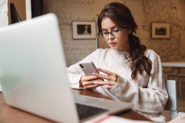 Kafede gözlük takan, dizüstü bilgisayar ve akıllı telefon kullanan beyaz bir kadın resmi.