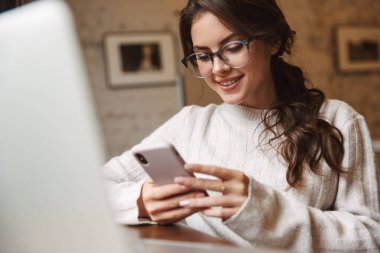 Kafede gözlük takan, dizüstü bilgisayar ve akıllı telefon kullanan beyaz bir kadın resmi.