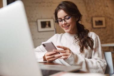 Kafede gözlük takan, dizüstü bilgisayar ve akıllı telefon kullanan beyaz bir kadın resmi.