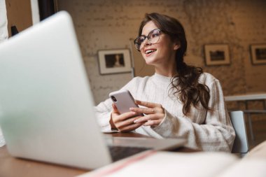 Kafede gözlük takan, dizüstü bilgisayar ve akıllı telefon kullanan beyaz bir kadın resmi.