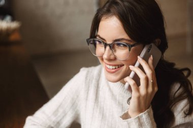 Kafede, gözlüklü, akıllı telefondan konuşan güzel beyaz bir kadın resmi.