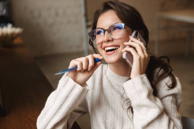 Kafede, gözlüklü, akıllı telefondan konuşan güzel beyaz bir kadın resmi.