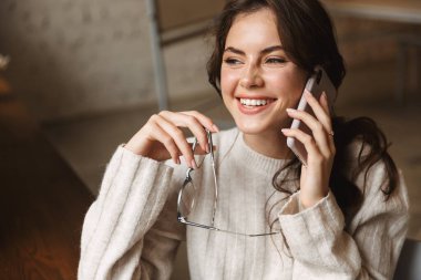 Kafede elinde gözlük olan ve akıllı telefondan konuşan güzel beyaz bir kadın resmi.