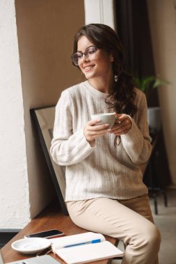 Kafe 'de uzun kahverengi saçlı, ders çalışan ve kahve içen güzel beyaz bir kadın resmi.