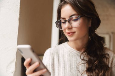 Kafede cep telefonu tutan uzun kahverengi saçlı güzel beyaz bir kadın resmi.