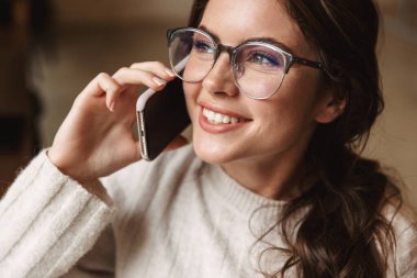 Kafede, gözlüklü, akıllı telefondan konuşan güzel beyaz bir kadın resmi.