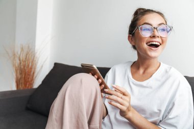Oturma odasındaki kanepede otururken gülümseyen ve cep telefonu kullanan gözlüklü heyecanlı bir kadın resmi