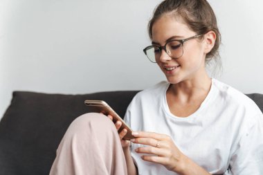 Oturma odasındaki kanepede otururken cep telefonunda gülümseyen ve daktilo kullanan gözlüklü hoş bir kadın resmi.